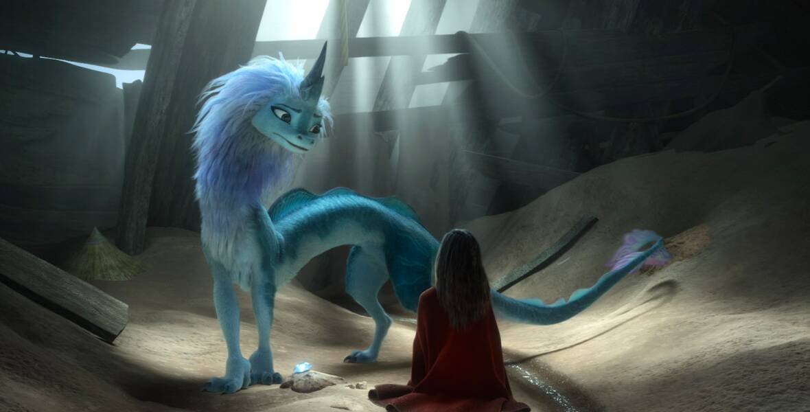 Sisu is in a space enclosed with wooden planks and sand on the bottom. Sitting cross-legged on the sand with her back toward the lens is Raya, who wears a red cloak. Sisu looks at Raya with a worrying expression, and a blue crystal lays on the sand between the two characters.