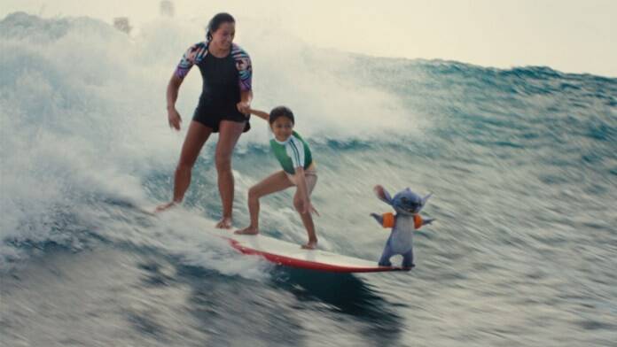 In a scene from Lilo & Stitch (2025), the sisters Nani (Sydney Elizebeth Agudong) and Lilo (Maia Kealoha) ride a surfboard on a tall ocean wave. At the front of the surfboard is Stitch (voiced by Chris Sanders).
