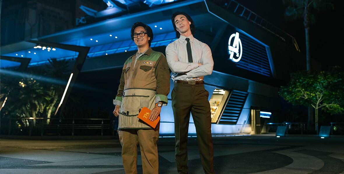 Two walk-around characters at Avengers Campus at Disney California Adventure Park are dressed as Loki and O.B. from Marvel Studios’ Loki. Loki is dressed in a light gray long sleeve collared shirt with a dark blue tie, gray trousers, and a black belt, and is standing with his arms crossed and a serious look on his face. O.B. is next to him, wearing a dark brown, light brown, and green patchwork jacket with brown slacks, glasses, and an apron; he is smiling while holding an orange book.